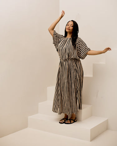 A woman modeling a black and beige vertically striped midi dress with wide short sleeves and a matching tie belt, standing on minimalist white steps.