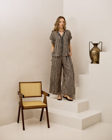 A woman models a matching two-piece striped matching set consisting of a short-sleeve top and wide-leg trousers, styled in a minimalist white room with a wooden cane-accent chair and a bronze vase on white steps.
