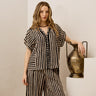 A woman models a matching two-piece set featuring a black and beige striped short-sleeve top and wide-leg pants, posed next to a decorative bronze vase on a white pedestal.