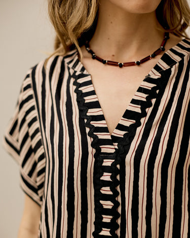 Close-up of a woman wearing a short-sleeve striped top with black and beige vertical stripes, a black scalloped V-neck trim, and a dark beaded necklace.