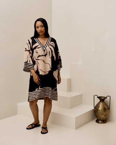 A woman models a black and beige abstract-patterned tunic dress with wide three-quarter sleeves and a V-neckline, standing on minimalist white steps next to a decorative bronze vase.