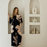 A woman modeling a black and beige floral print matching set in a minimalist white room with arched recessed shelves.