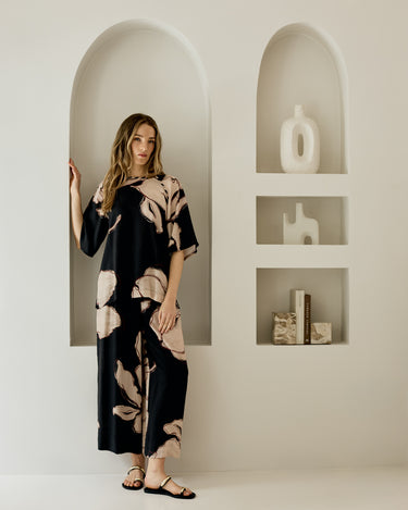 A woman modeling a black and beige floral print matching set in a minimalist white room with arched recessed shelves.