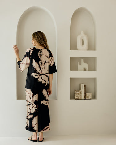 A woman stands in front of a white wall with two recessed arched shelves, wearing a black matching set with a beige floral pattern. She is seen from behind, looking toward the left shelf.