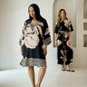 A woman models a loose black tunic dress with a beige floral print and striped hem in a bright studio with arched alcoves in the background. Another woman stands blurred in the back wearing a matching set.