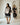 A woman models a loose black tunic dress with a beige floral print and striped hem in a bright studio with arched alcoves in the background. Another woman stands blurred in the back wearing a matching set.