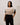 A woman modeling a color-block short-sleeve blouse with a white upper yoke, beige center panel, and black sleeves, paired with black pants against a plain light background.