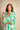A woman wearing a green and white patterned button-up shirt dress, posing against a textured beige background.