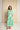 A woman modeling a long-sleeve maxi dress with a bold green and white geometric pattern, standing in front of a large beige textured backdrop.