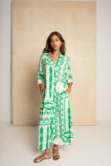 A woman modeling a long-sleeve maxi dress with a bold green and white geometric pattern, standing in front of a large beige textured backdrop.