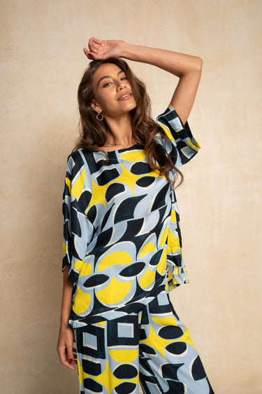 A woman modeling a matching two-piece set featuring a bold abstract pattern in yellow, navy blue, and light blue, posed against a textured beige background.