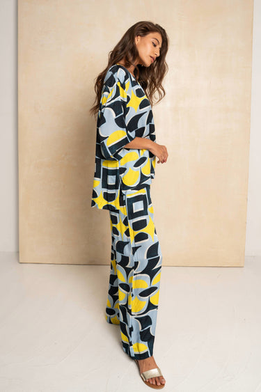 A woman modeling a matching two-piece set with a bold abstract geometric pattern in yellow, navy, and light blue, standing against a beige textured wall.