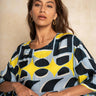A woman wearing a loose-fitting short-sleeve top with a bold abstract geometric pattern in light blue, yellow, and navy, posed against a neutral beige textured background.