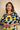 A woman wearing a loose-fitting short-sleeve top with a bold abstract geometric pattern in light blue, yellow, and navy, posed against a neutral beige textured background.